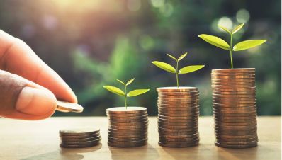 A hand places a coin on a small stack of coins. Three taller stacks, each with a growing plant, symbolize financial growth and investment, set against a blurred natural background.
