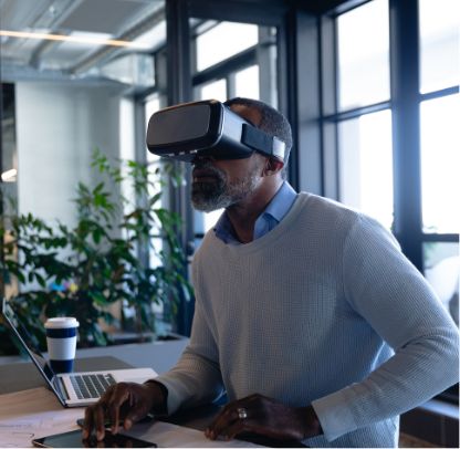 A man wearing a virtual reality headset sits at a desk in a modern office, with a laptop, papers, and a coffee cup nearby. Large windows and green plants are visible in the background.