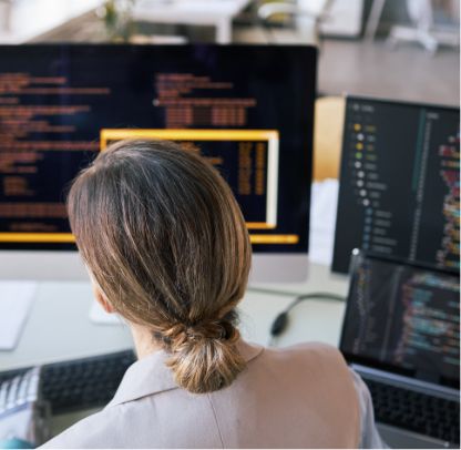 A person with light brown hair tied in a low bun is sitting at a desk, working on multiple computer screens displaying lines of code in a modern office setting.