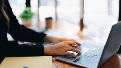A person types on a laptop at a wooden desk with a notebook, papers, and a blurred green plant and coffee cup in the background.