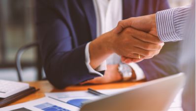Two people in business attire shake hands at a desk with documents, a laptop, and a calculator, suggesting a successful business agreement or partnership.