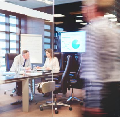 Two people sit at a conference table in a modern office, discussing work with papers in front of them. A presentation screen shows a pie chart. A blurred person walks past the glass door in the foreground.
