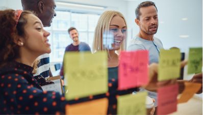A group of five people in an office collaborates, smiling and discussing colorful sticky notes arranged on a glass wall during a team meeting.
